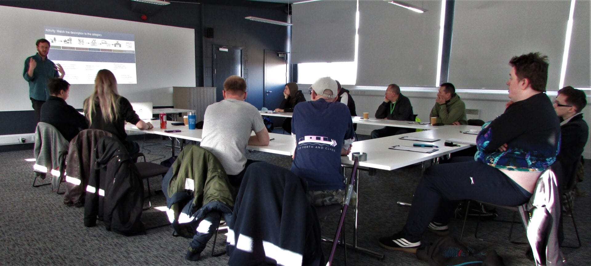 A Keep Scotland Beautiful trainer stands at the front of a classroom and shows a slide on a projector screen. 10 students sit at their desks while listening to him,