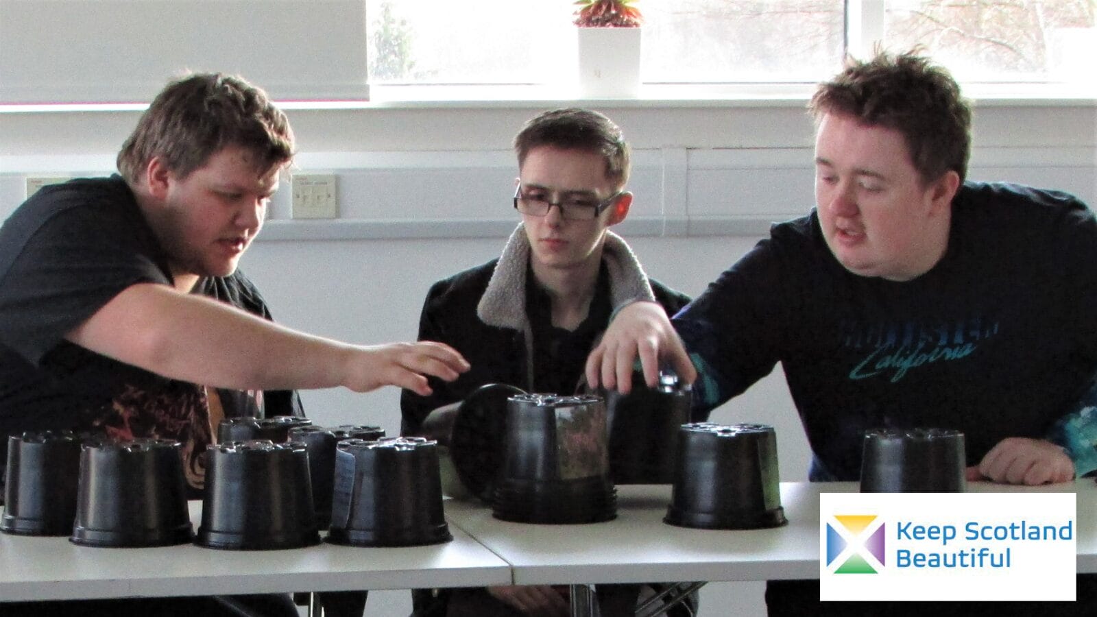 Three students in a classroom sitting at a desk that has several black plastic upside down plant pots on it. They are having a discussion about the plant pots are their positioning on the table.