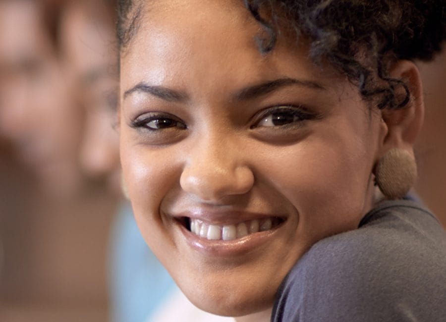 A young woman sits, smiling over her shoulder. She's sitting next to other people, who are blurred into the background. She signifies a person whom the SCQF Framework supports.