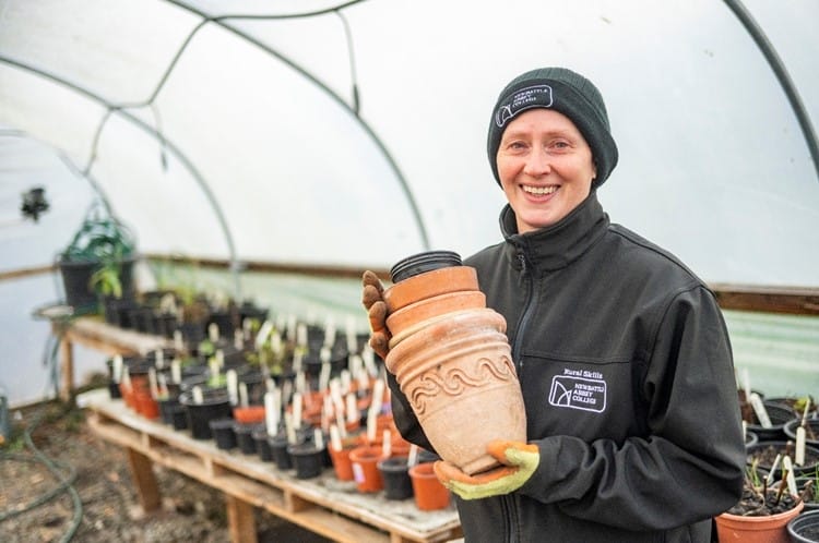 Lee Ryan, at student of Newbattle Abbey College, holds up various ceramic pots. She is standing inside a polytunnel and there are lots of plants on the planting benches behind her.