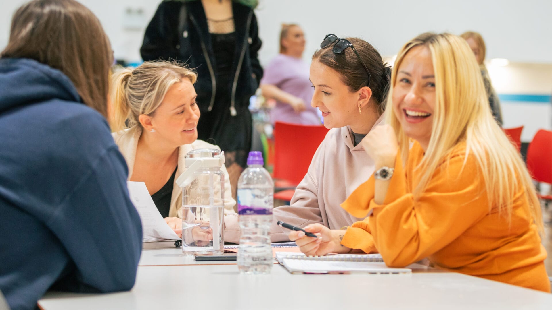 Four women chatting at a table while writing in notepads. One of the women is smiling at the camera