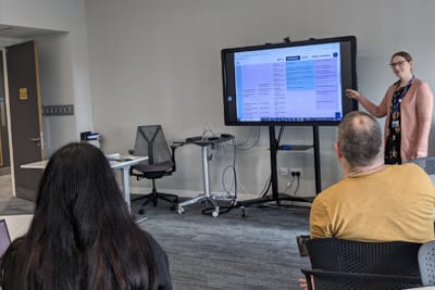 Gillian WIlson stands next to a television screen, which is on a tall stand. Adults are sitting around, watching her presentation. The screen features the SCQF diagram
