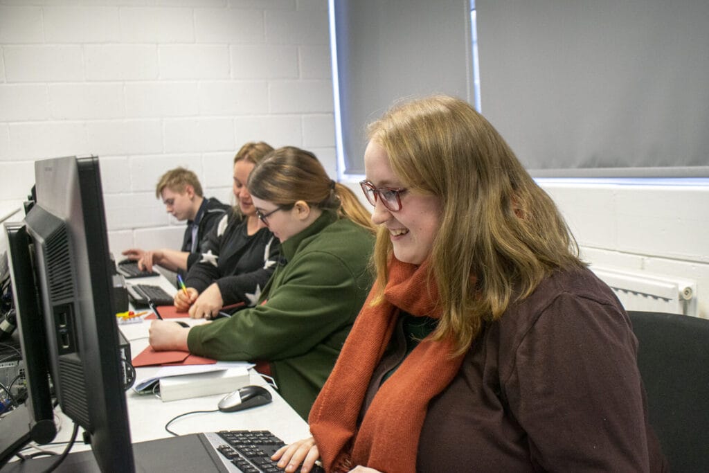 Four people are sitting at a long desk, each using a desktop computer. Two of them are chatting while writing on red paper folders.