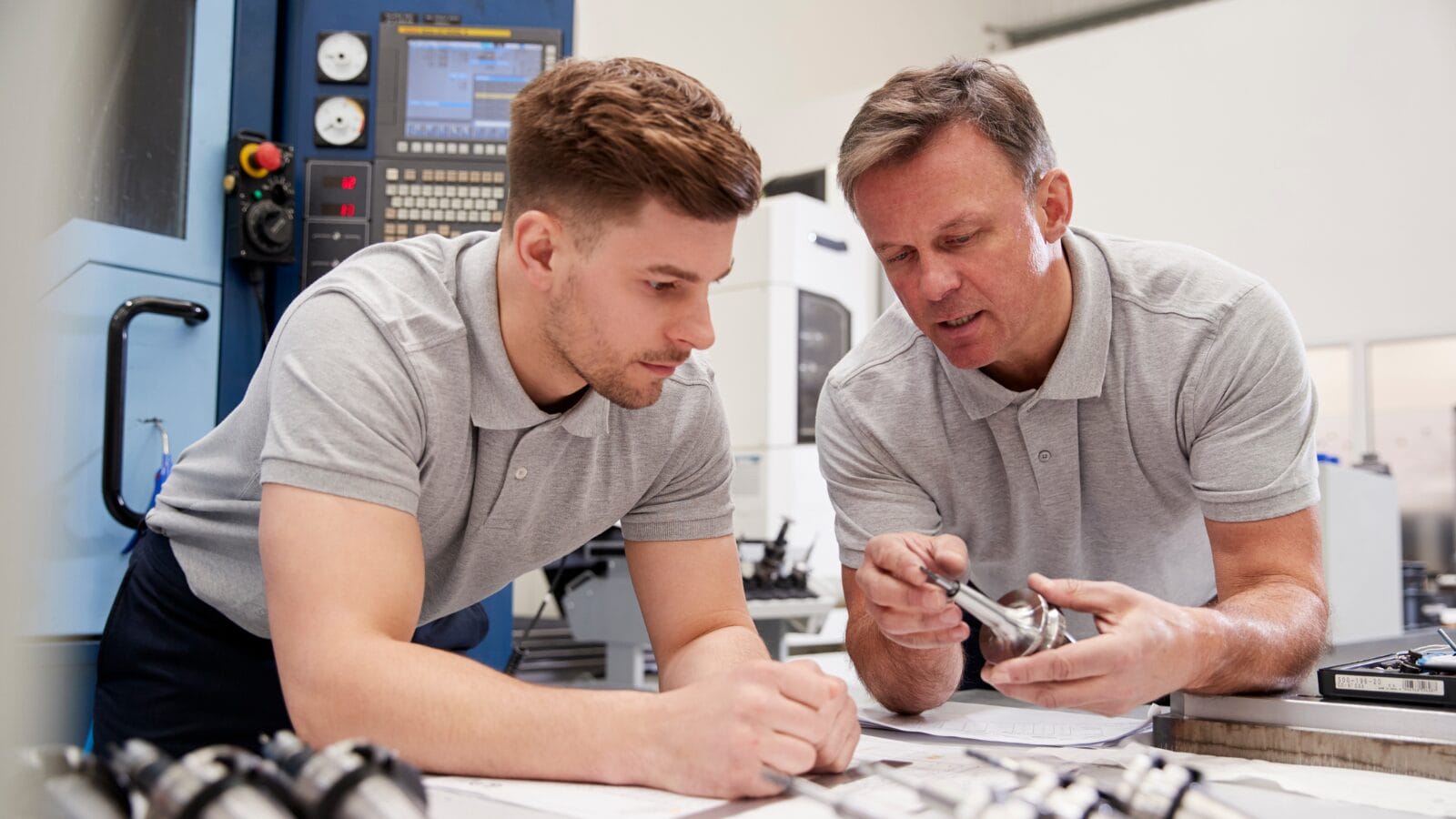 A younger and old man in grey polo shirts, leaning over a table within a factor setting. The older man is showing the younger man how a tool works. The younger man signifies an apprentice.