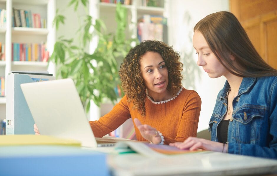 A younger and older woman sit at a desk. The older woman is talking to the younger woman. There is a laptop on the deck.