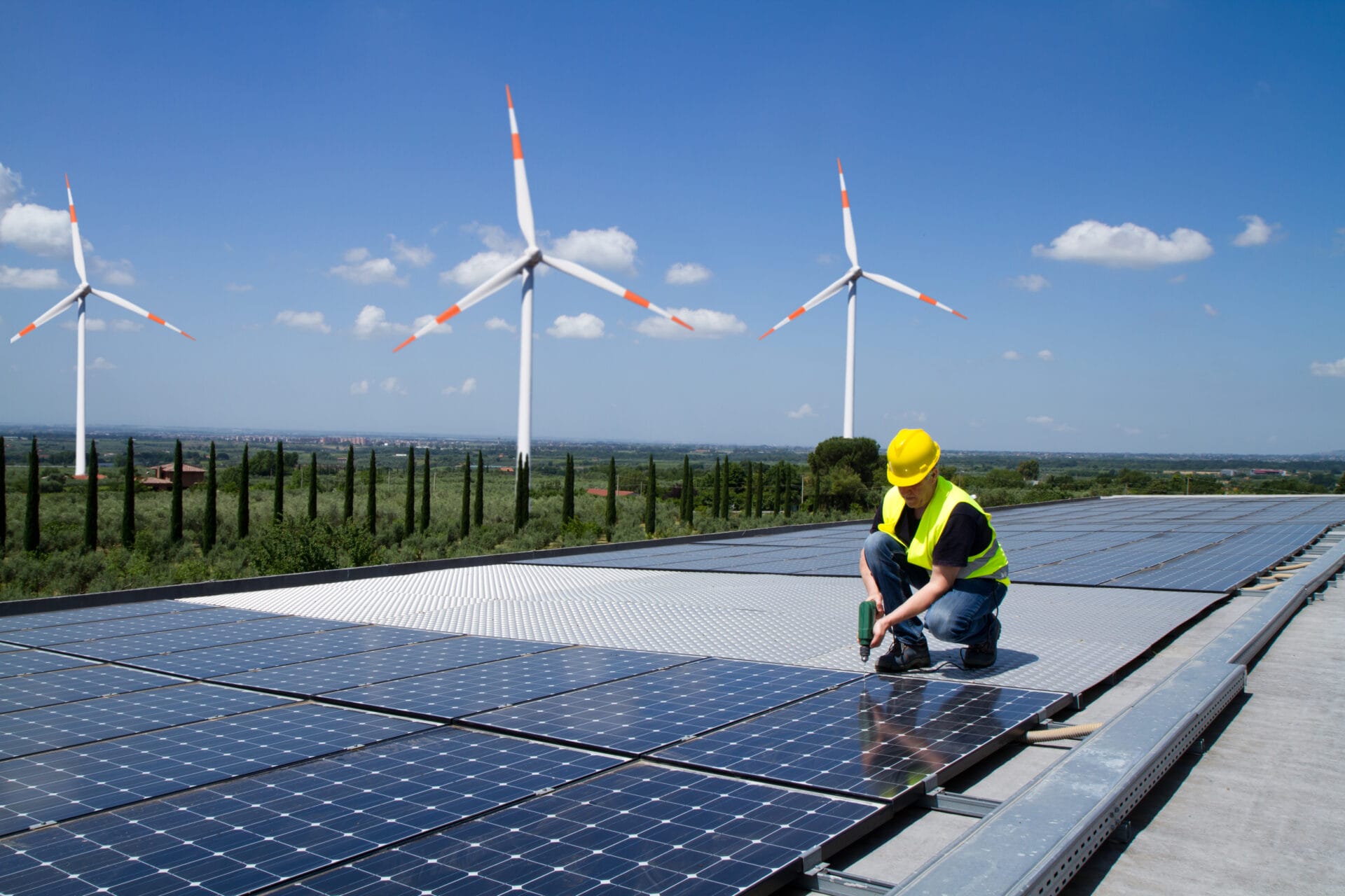 A man is at a renewable energy farm. There's blue skies in the background and 3 wind turbines in a field. In front of the field lie several solar panels on the ground. The man, who is in a hard hat and wearing a hi-vis vest is kneeling over a solar panel and working on it with tools. The photo represents the various industries the SCQF supports.