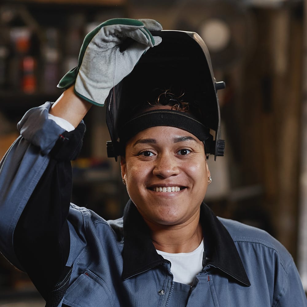 A woman wearing blue overalls and a welder's safety guard on her head. She signifies the range of learners and employees that the SCQF supports.