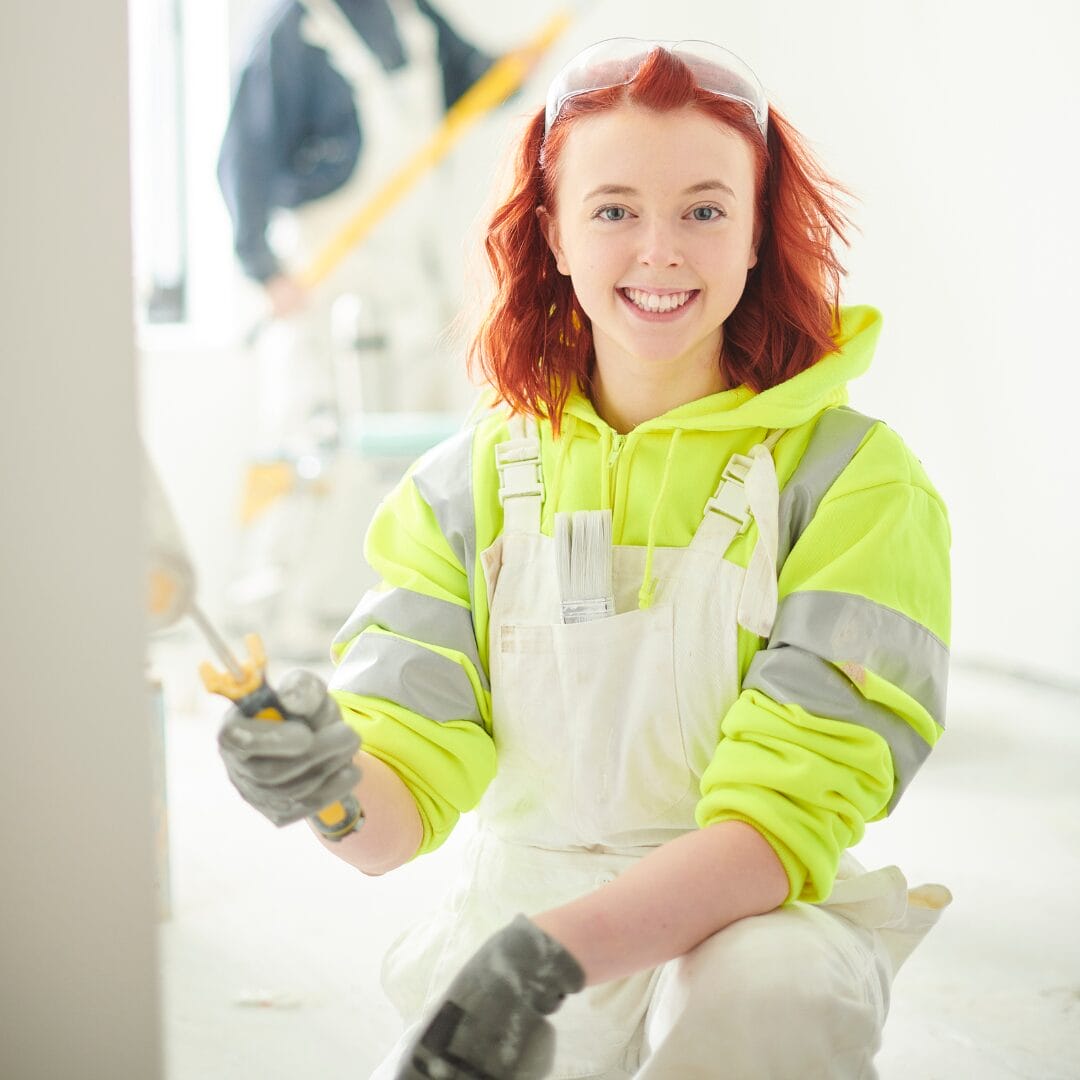 A young women in fluorescent workwear and overalls with safety goggles sitting on top of her head. She's holding a paint roller and painting a wall white. She signifies the range of learners and employees that the SCQF supports.