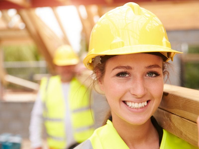 Female apprentice wearing hard hat and carrying timber
