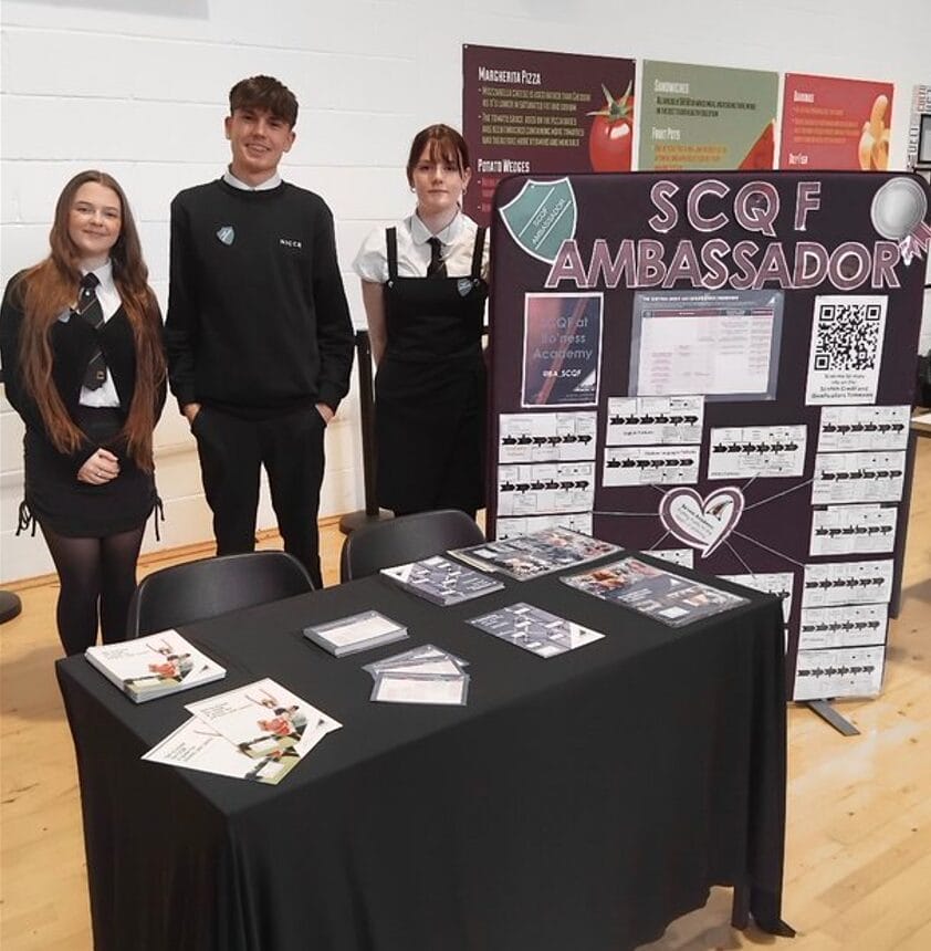Three Bo'Ness Academy SCQF Ambassadors standing behind a desk that features SCQF leaflets for parents' night. Their SCQF board is to the right, showing the different pathways on offer at the school
