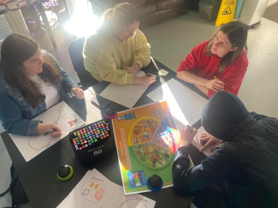 4 people sit at a table; 3 are writing on paper, while another in a red jumper is talking to the group