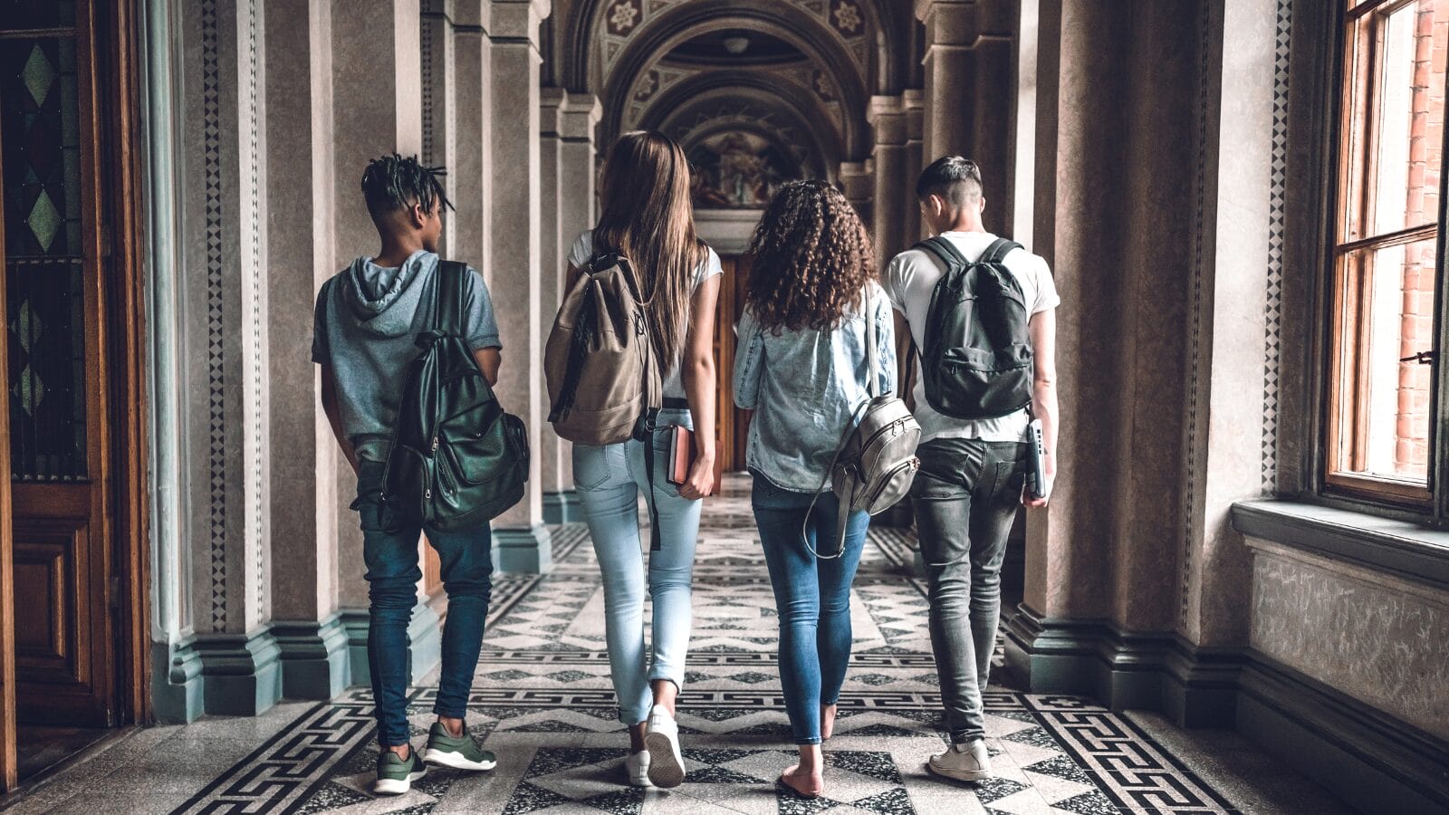 Four university students walking down a university corridor, with their backs facing the camera. They are carrying backpacks.