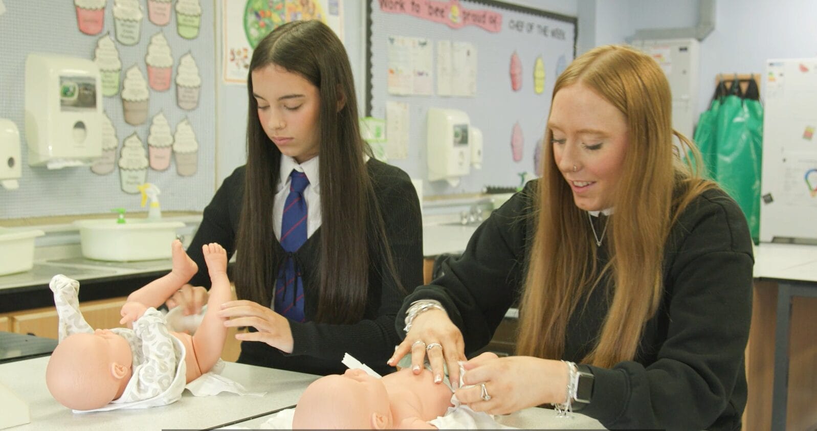 2 female pupils wearing Craigmount High School uniform, at a desk. Both have a plastic baby doll and they're putting nappies on the doll.