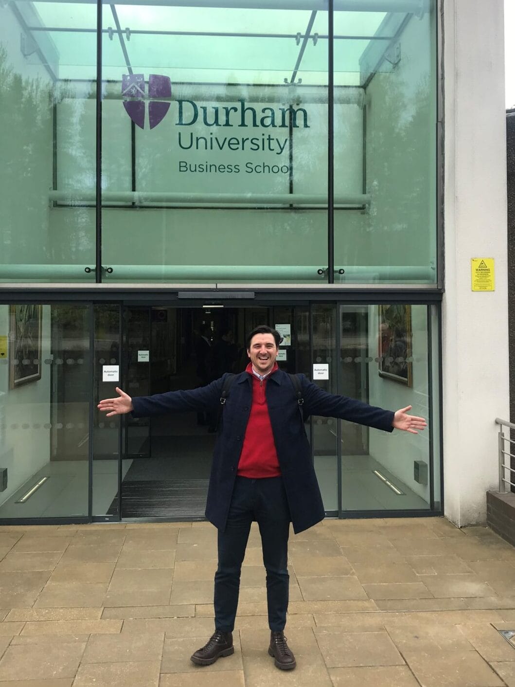 David Balfour standing with his arms outstretched and smiling. He's outside the entrance to Durham University Business School