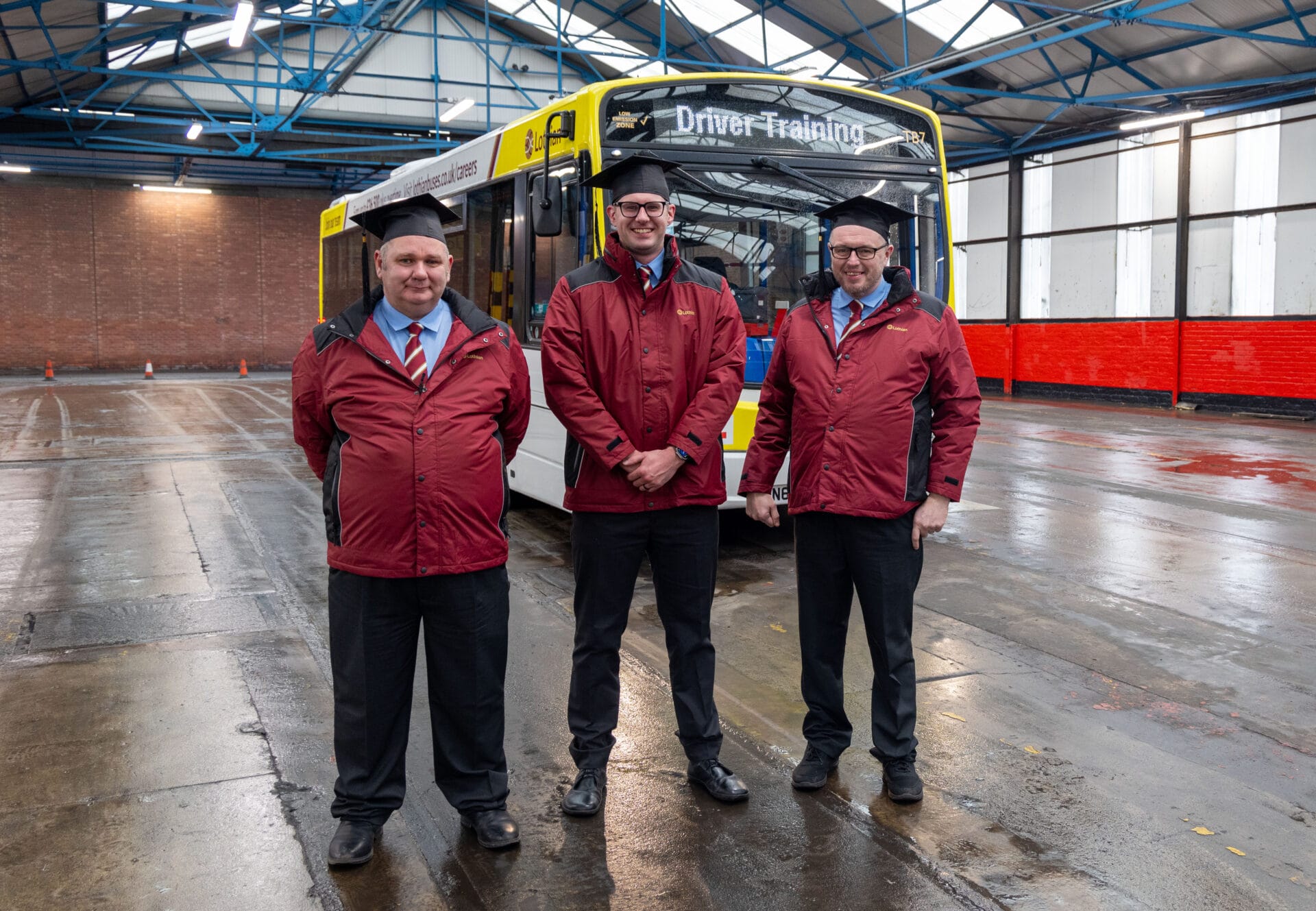 Three men wearing red Lothian Buses jackets, black trousers, and ties stand in front of a yellow and white bus marked "Driver Training" inside a bus depot. They are smiling and wearing black graduation caps, symbolizing the completion of their SCQF level 6 bus driver qualification. The depot has a high ceiling with blue steel beams, and the floor appears wet.