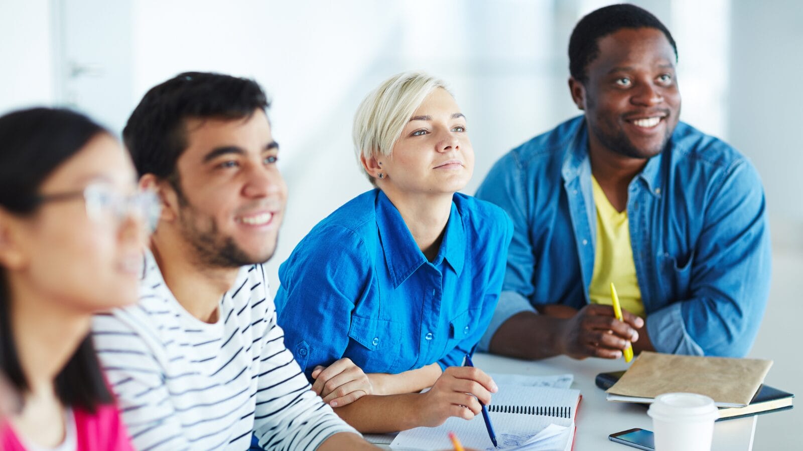 A group of four adult learners sitting at a table in a bright classroom, attentively listening to something off-camera. They have notebooks, pens, and a coffee cup in front of them, appearing engaged and focused.