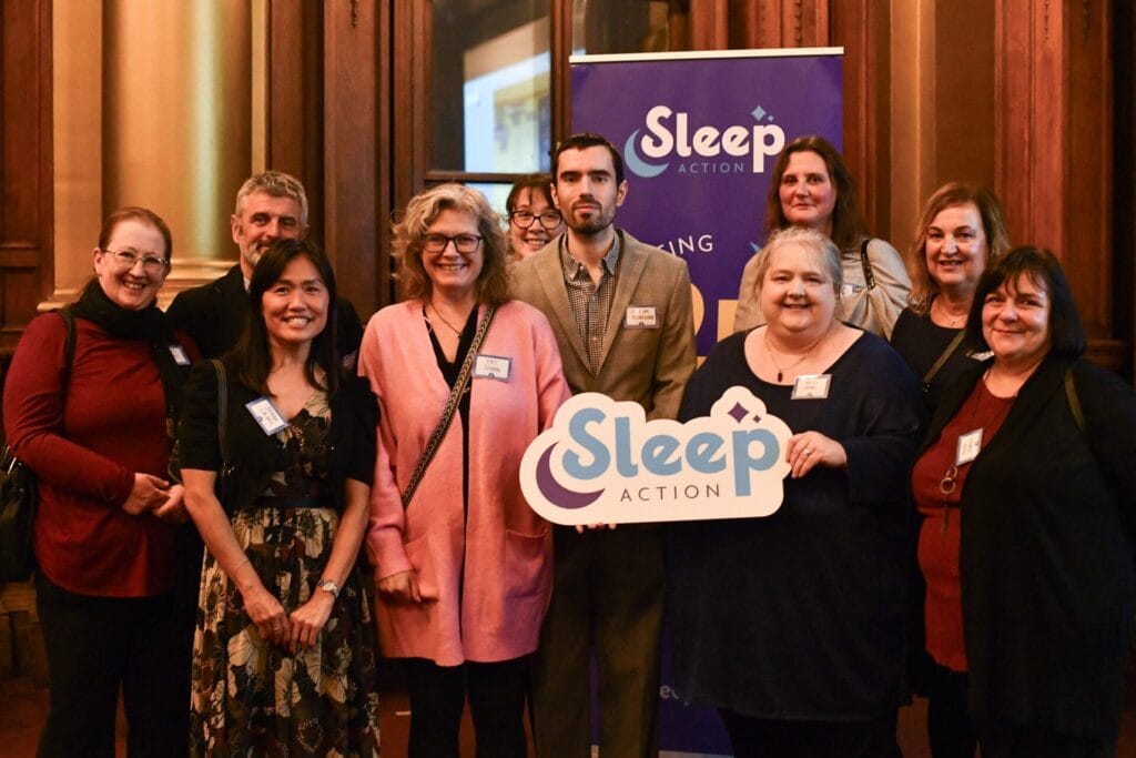 A group of 12 people smiling and posing for a photo at an indoor event. They are standing in front of a purple banner that reads “Sleep Action.” One person in the front is holding a large Sleep Action sign. The group includes men and women of various ages, dressed in smart casual attire.