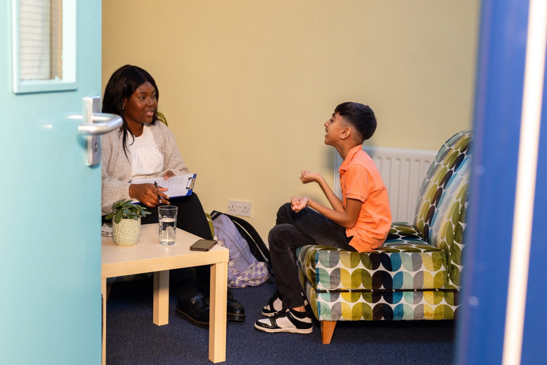 A young boy sits at a table with a young female therapist in a community centre. The boy is engaged in a supportive counselling session, actively participating in the discussion. The therapist is warm and approachable, creating a nurturing environment. The room is brightly lit and welcoming, with comfortable seating, reflecting a safe space for sharing and healing.