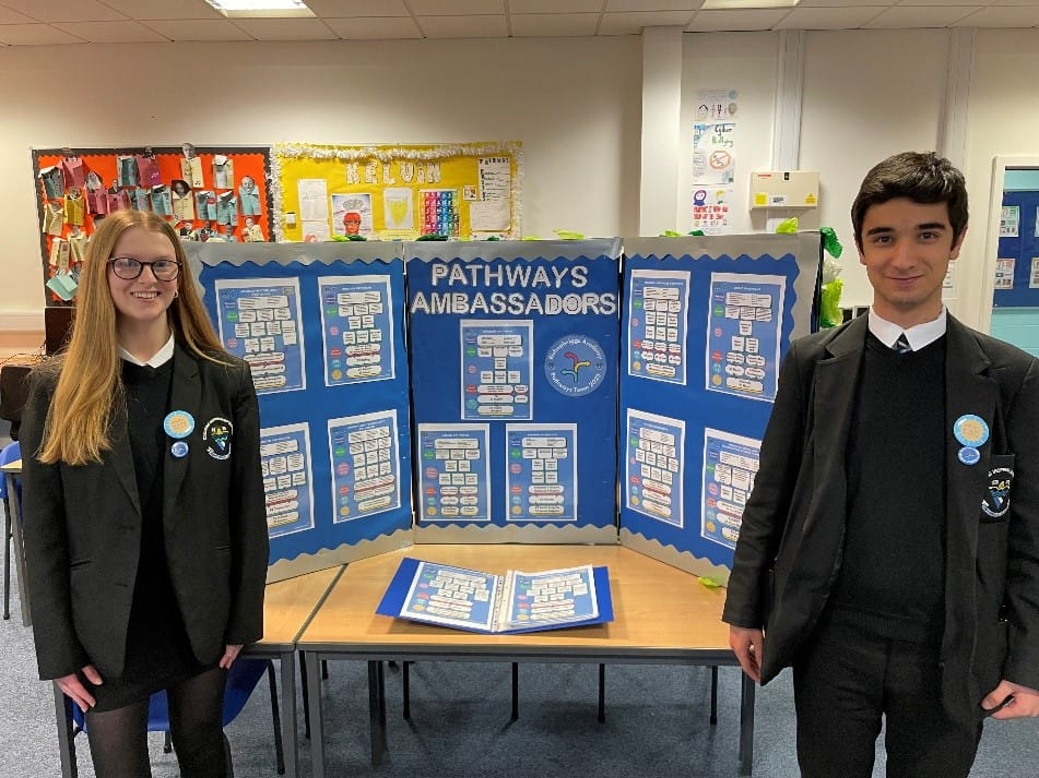 Two SCQF Ambassadors from Bishopbriggs Academy stand at either side of their pathways stand, which is sitting on a table