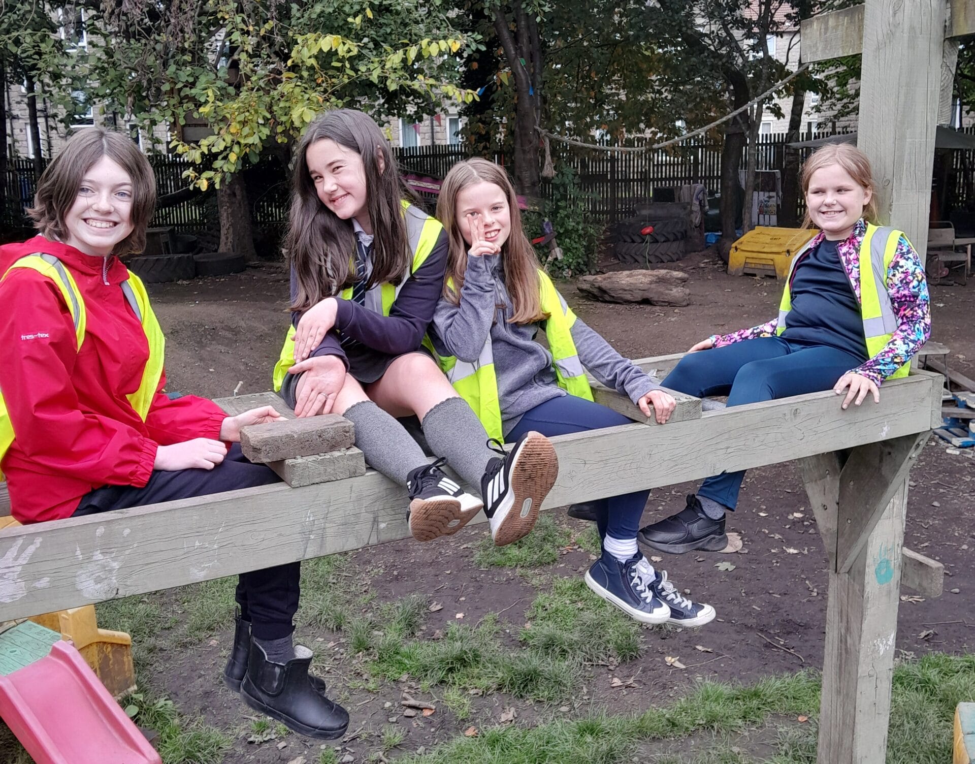 Four children wearing bright yellow safety vests sit and smile on a wooden play structure at Baltic Street Adventure Playground, surrounded by trees and outdoor play equipment