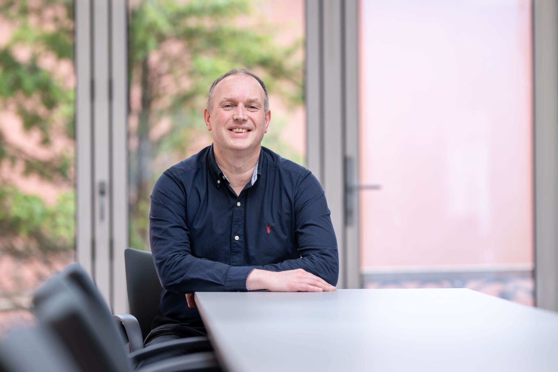 Ewen Hay sits casually at a desk, smiling, in a navy shirt. A closed window is behind him
