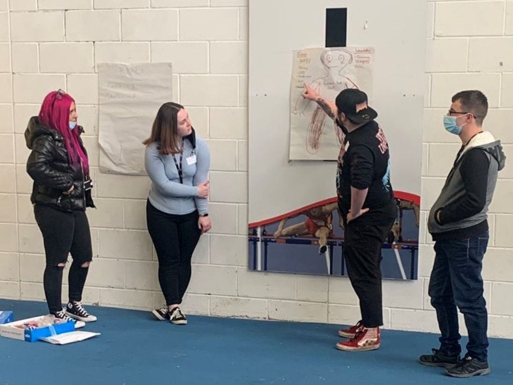 A group of young people in room, looking at a board which features a drawing of a person. One young man is pointing at the drawing.