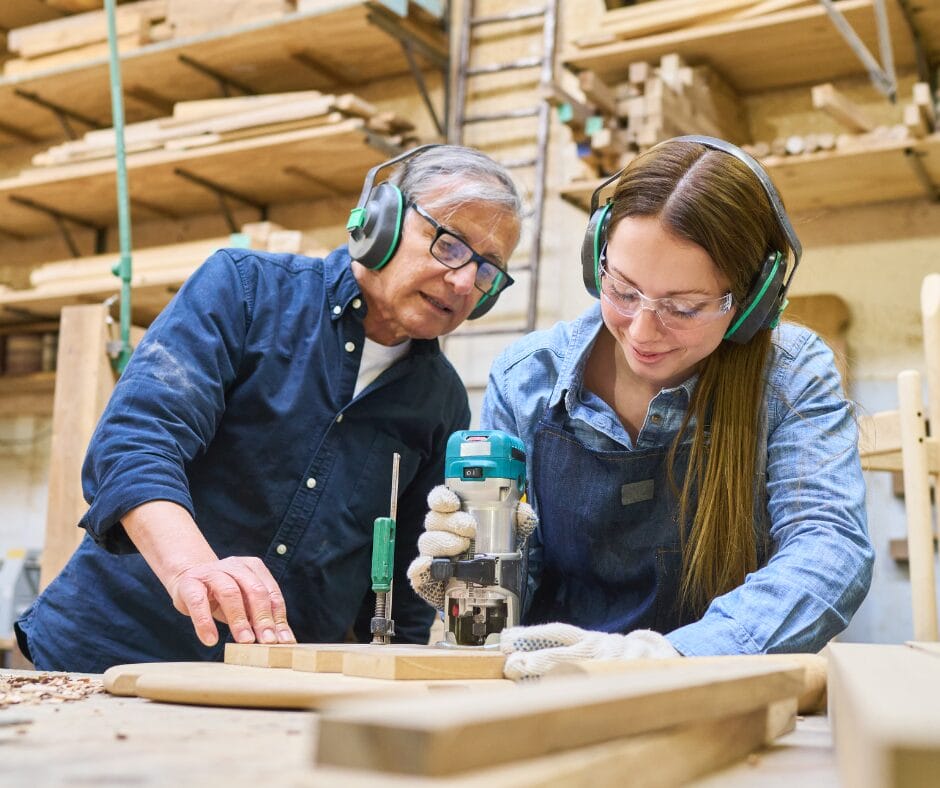 An older man supervises a young woman in a carpentry workshop as she uses a tool on a piece of wood