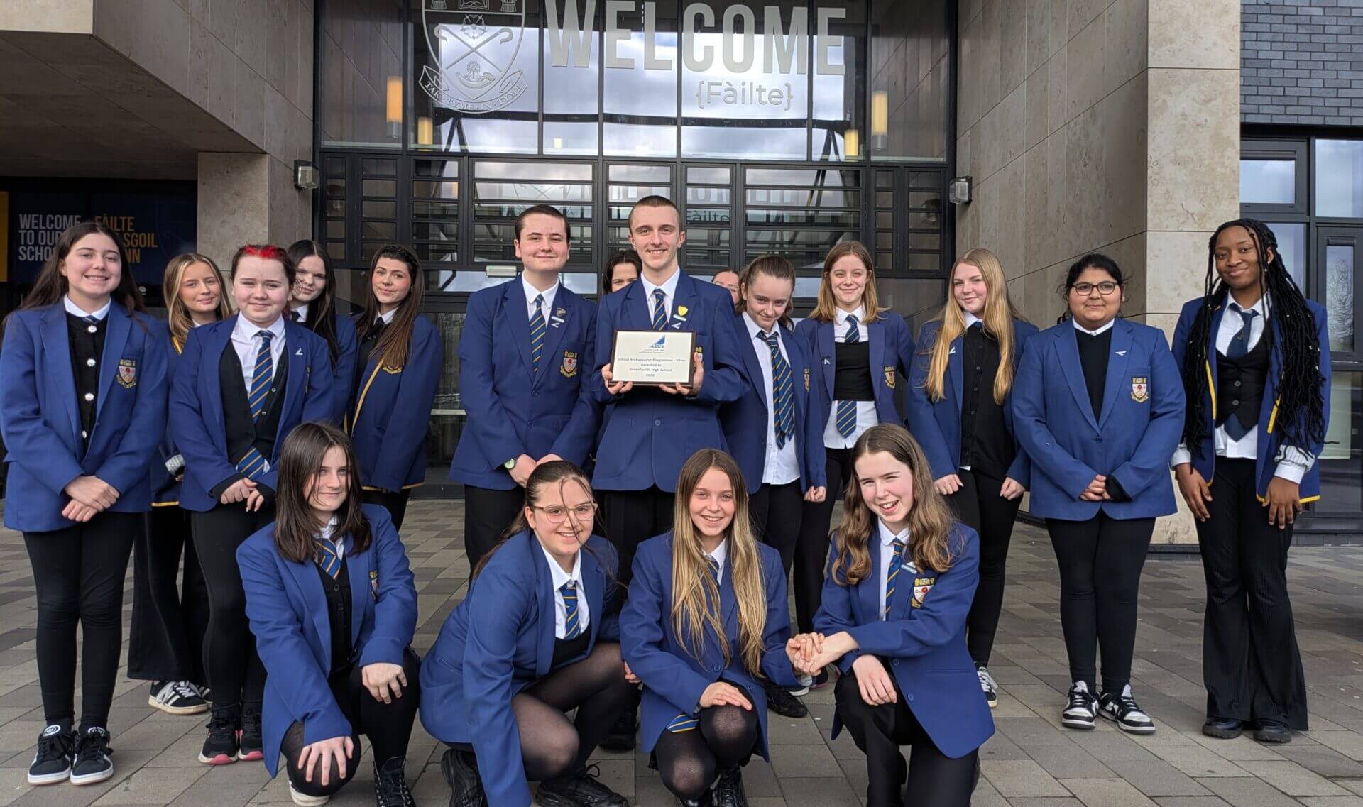 Greenfaulds High School pupils in blue school uniforms pose outside their school entrance beneath a large “Welcome (Fàilte)” sign. Two students at the centre hold a silver SCQF Ambassador plaque while classmates stand and kneel around them, smiling for the photo.
