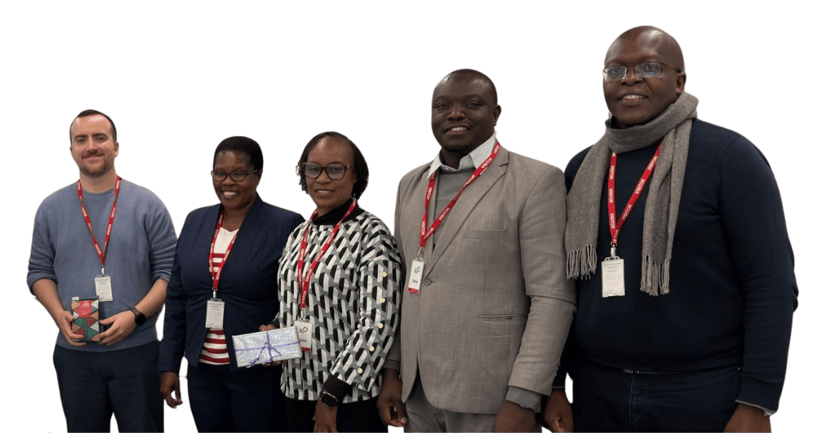 Paul White, SCQFP Lead for International, standing on the left with a group of four visitors from Kenya during a study visit to Forth Valley College. All are wearing conference lanyards and smiling at the camera, with two members of the group holding wrapped gift boxes. The group is dressed in smart-casual to business attire
